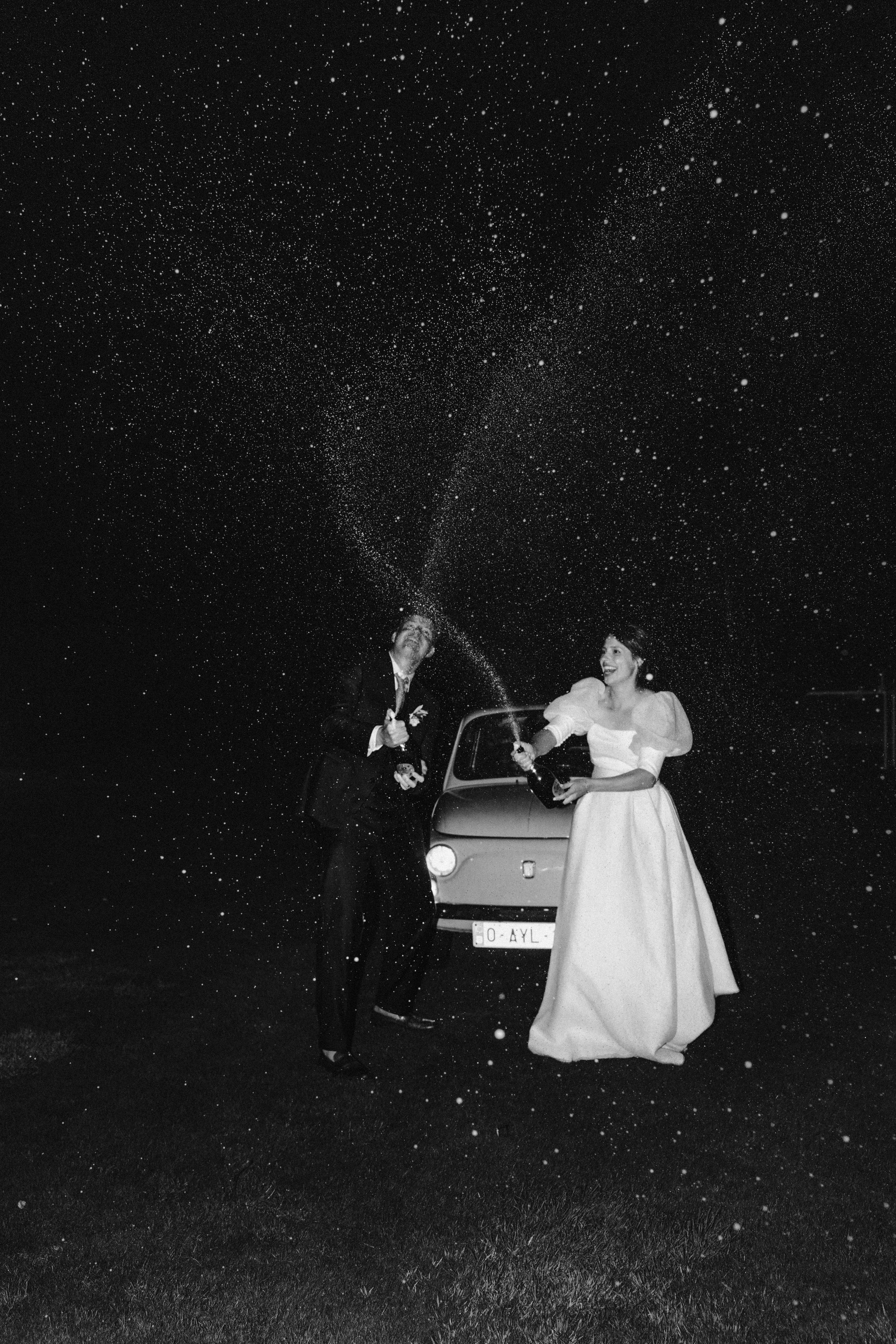 Two just married people standing next to a car under a starry night sky.