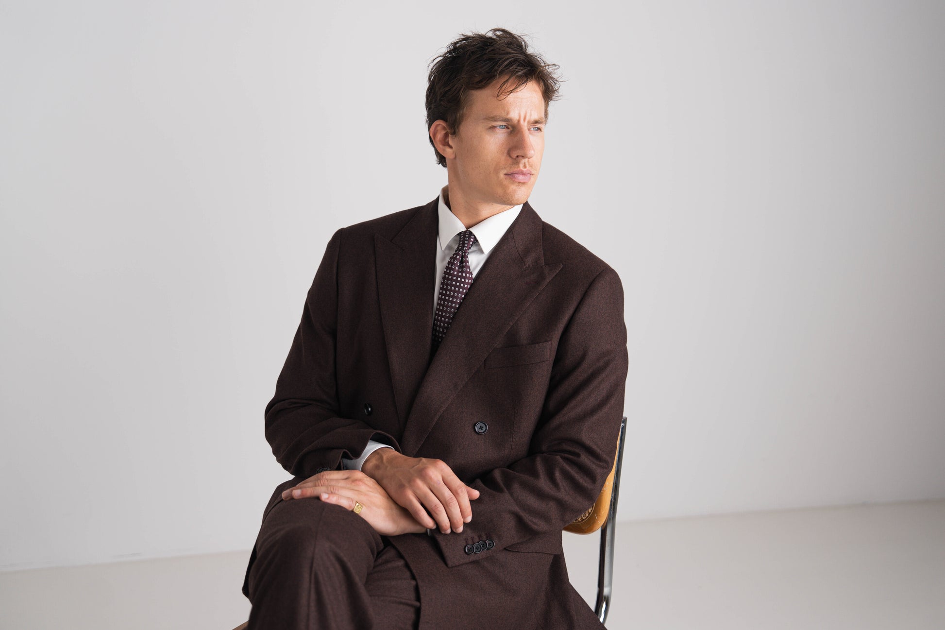 Man in a brown suit sitting on a chair against a plain background